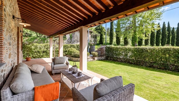 Eine überdachte Terrasse mit Korbmöbeln, einer Steinwand und Blick auf einen gepflegten Garten.
