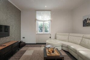 Una sala de estar moderna con un televisor de pantalla plana, un sofá de cuero, una mesa de centro con pasteles y una ventana con vistas a zonas verdes.