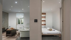 A modern hotel room with a bed, a sofa, a small table with a tray of pastries, a window with curtains, and a wall-mounted light fixture. - Quiet City Apartment Dexa, Rijeka, Croatia (Rijeka)