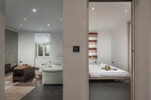 Ein modernes Hotelzimmer mit Bett, einer Couch, einem kleinen Tisch mit einer Etagere voller Gebäck, einem Fenster mit Vorhängen und einer Wandleuchte.