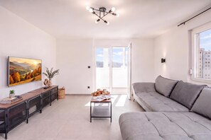 Ein modernes Wohnzimmer mit einem grauen Sofa, einem Couchtisch mit Büchern und einer Muffelplatte, einem Landschaftsbild und einer Deckenleuchte.