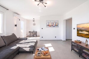A modern living room with a grey sectional sofa, a coffee table with a tray of muffins, a painting of sailboats, and a television displaying a landscape. - City Seaview Apartment Vigo, Rijeka, Croatia (Rijeka)