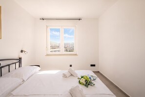 A bedroom with a bed, a window with a city view, a bedside table, and a wall-mounted light fixture. - City Seaview Apartment Vigo, Rijeka, Croatia (Rijeka)