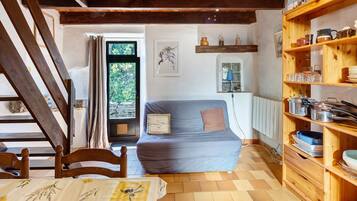 A cozy room with a wooden staircase, a sofa, a dining table with a floral tablecloth, and a wooden shelf with kitchenware.