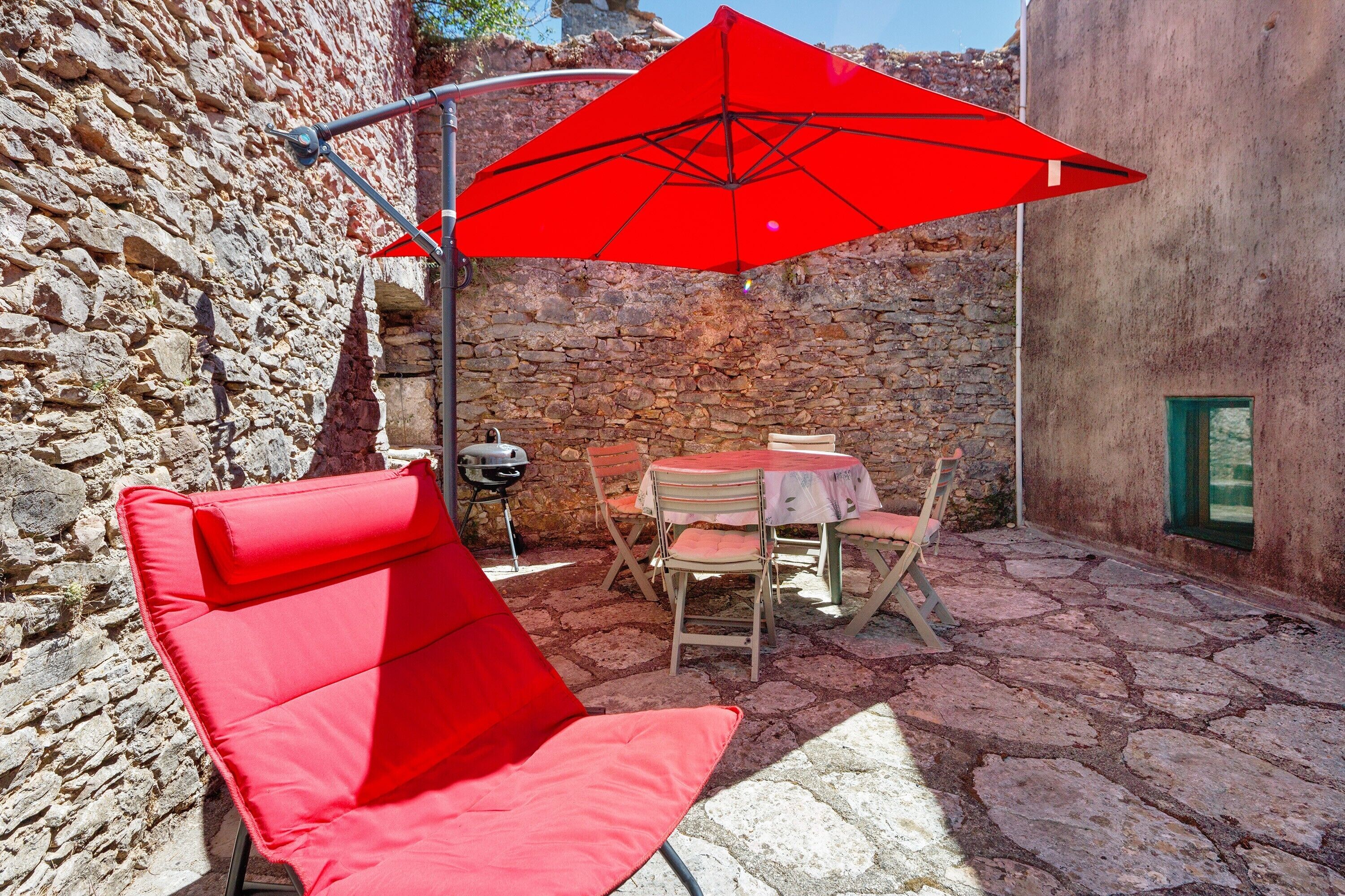 Une terrasse en pierre avec un parasol rouge, une table avec des chaises et un fauteuil de jardin rouge.