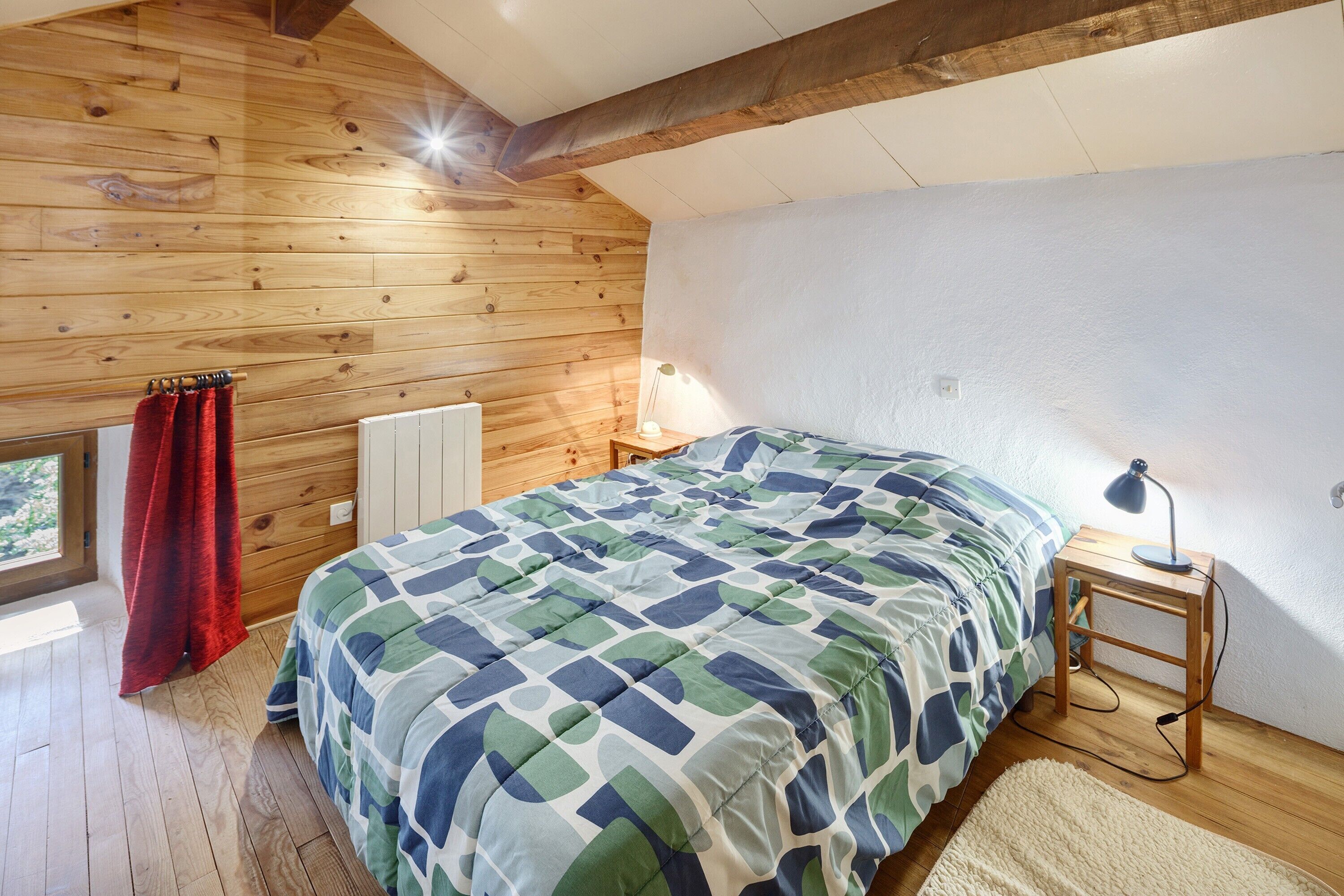 Een slaapkamer met een houten plafond, een bed met een sprei met een geometrisch patroon, een nachtkastje met een lamp en een raam met gordijnen.