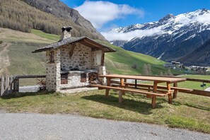 Een rustiek stenen gebouw met een houten picknicktafel, omgeven door een gazon en een berg op de achtergrond.
