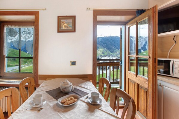 Ein Essbereich mit Tisch für zwei, Blick auf eine Berglandschaft durch große Fenster und eine Küchenzeile mit einer Mikrowelle.