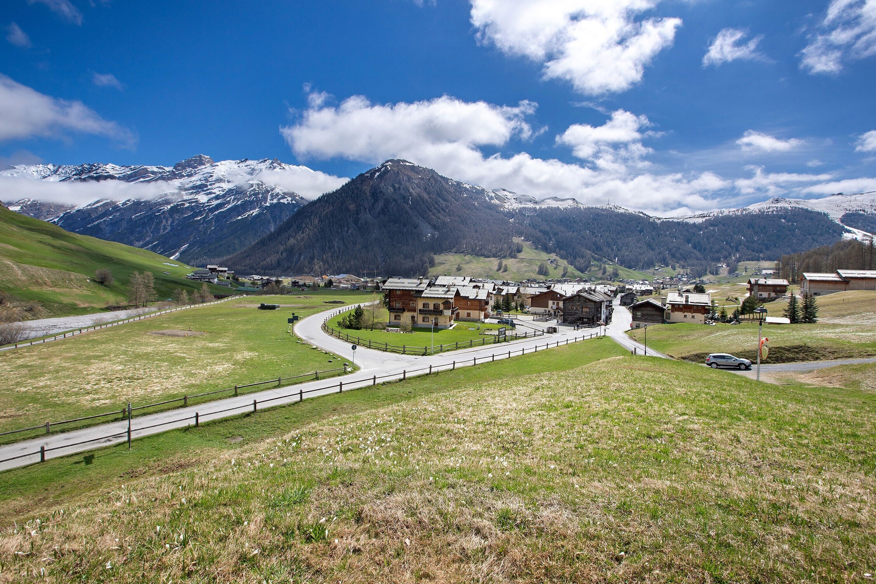 A mountainous village with traditional buildings, a winding road, and snow-capped peaks in the background.