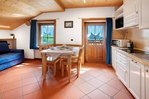 Een gezellige kamer met een eettafel, stoelen, een bank, een keukenhoek met kastjes en een magnetron, en een raam met gordijnen.