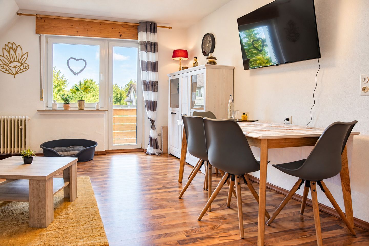 A modern living room with a wooden floor, a dining table with chairs, a TV mounted on the wall, and a view of greenery outside.