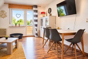 A modern living room with a wooden floor, a dining table with chairs, a TV mounted on the wall, and a view of greenery outside. - Fewo Renauweg - Gemütliche Ferienwohnung in Ruhiger Lage (Winterberg)