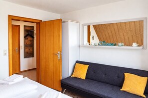 Une pièce comprenant un lit, un canapé gris avec des coussins jaunes, une armoire en bois et une étagère ornée d’objets décoratifs.