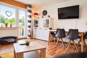 Un salon moderne avec un plancher en bois, un coin repas avec une table et des chaises, un téléviseur fixé au mur et un balcon agrémenté de plantes et d’une décoration en forme de cœur.