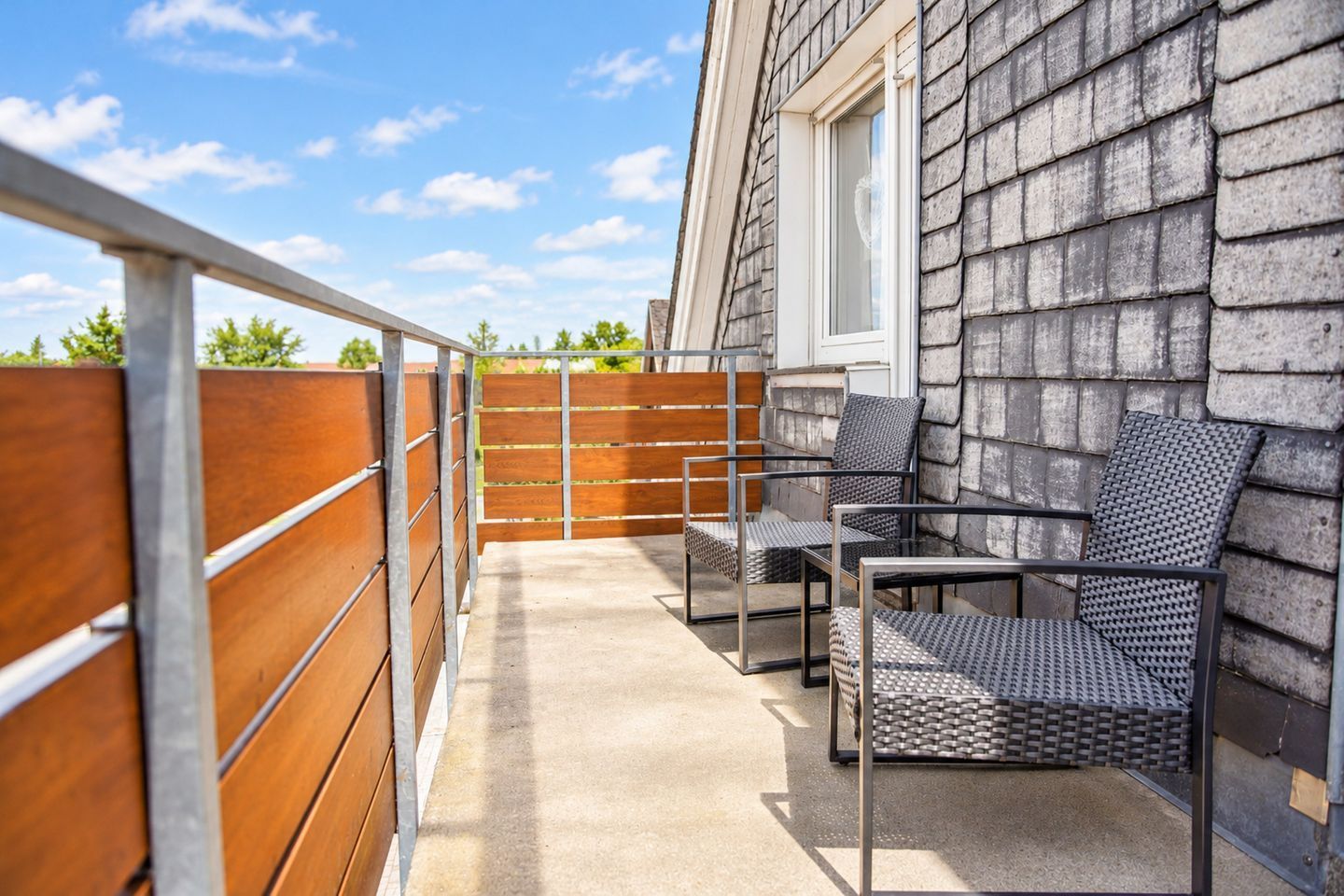 An outdoor patio area with a concrete floor, metal railing, and a wooden fence. There are two metal chairs with woven seats and backs.