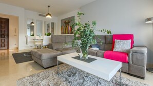 Una sala moderna con un sofá gris, una mesa de centro blanca y una planta. Al fondo, se aprecia un comedor con una mesa y sillas blancas.