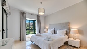 Ein modernes Schlafzimmer mit einem großen Bett, Nachttischlampen, Blick durch das Fenster und einem kleinen Nachttisch.