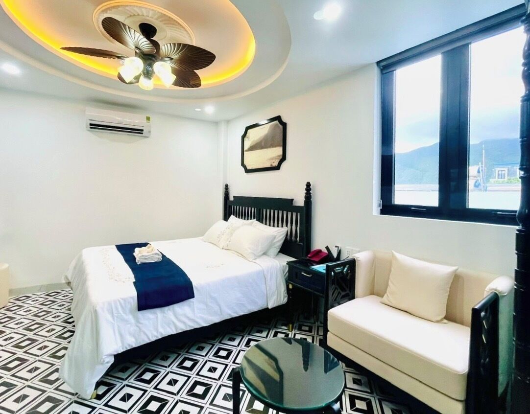 A hotel room with a bed, a sofa, a small table, a ceiling fan, and a window with a view.