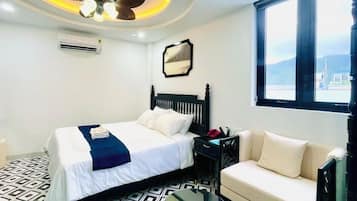 A hotel room with a bed, a sofa, a small table, a ceiling fan, and a window with a view.