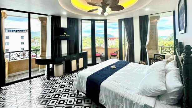 Ein Hotelzimmer mit Bett, Fernseher, Deckenventilator und Balkon mit Blick auf Gebäude und Grünflächen.
