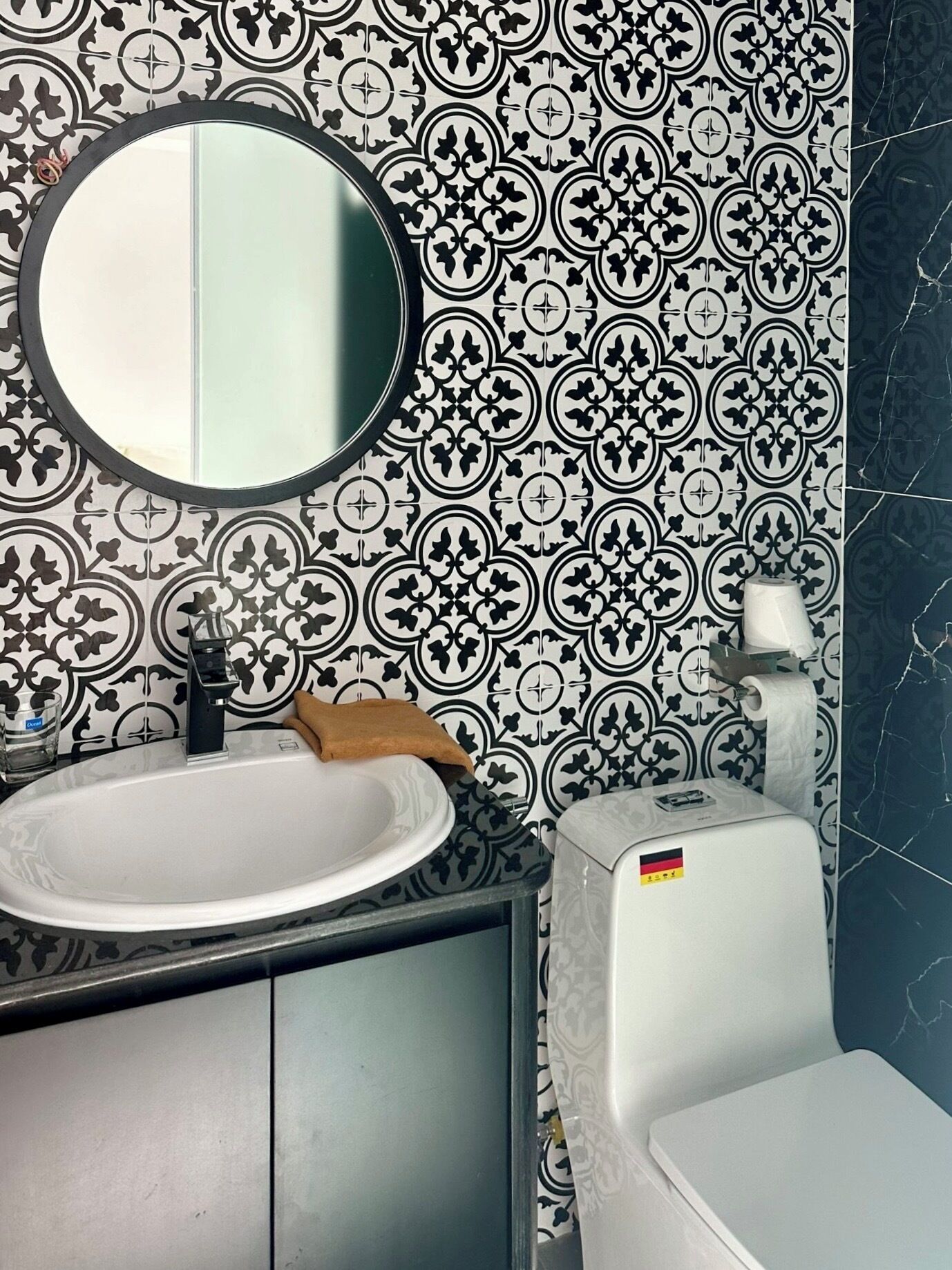 A bathroom with a white sink, a round mirror, a toilet, and patterned tiles.