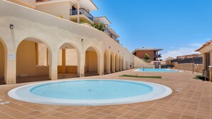 Un área de piscina con suelo de baldosas, aberturas en arco y un edificio con balcones.