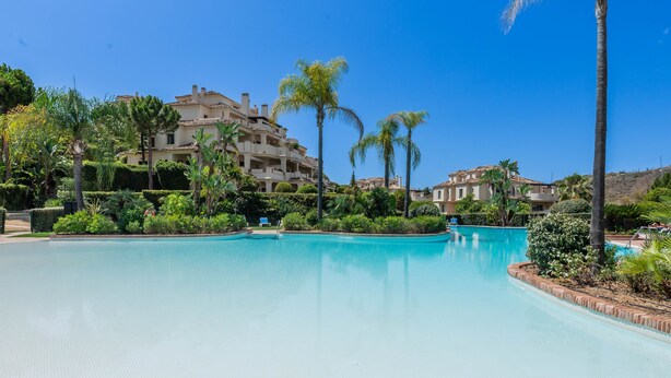 Une piscine entourée d’une végétation luxuriante et un grand bâtiment doté de nombreux balcons.