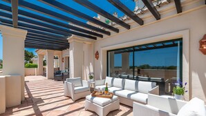 Une terrasse extérieure avec des meubles blancs, une table en bois sur laquelle se trouve une bouteille, et une grande fenêtre.