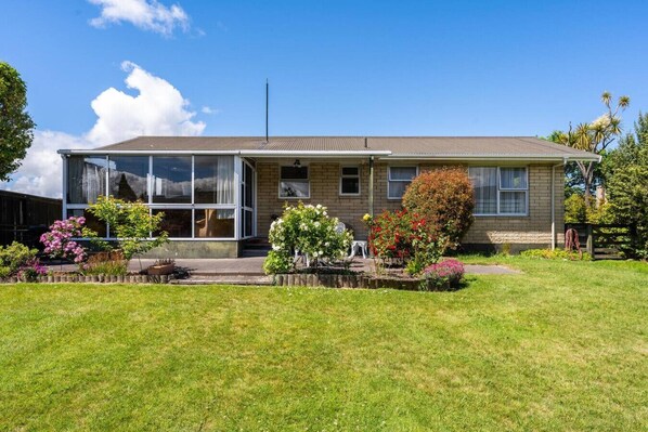 A two-story house with a large front window, a covered porch, and a well-maintained lawn with various flowering plants. - Classic Bach with Wifi  and pets (Taupō)