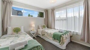 A hotel room with two single beds, a small table with a lamp, and large windows with curtains.