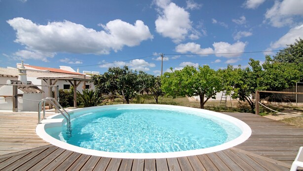 Une terrasse en bois avec une piscine ronde, une clôture blanche et, au fond, un bâtiment dont le toit est recouvert de tuiles rouges.