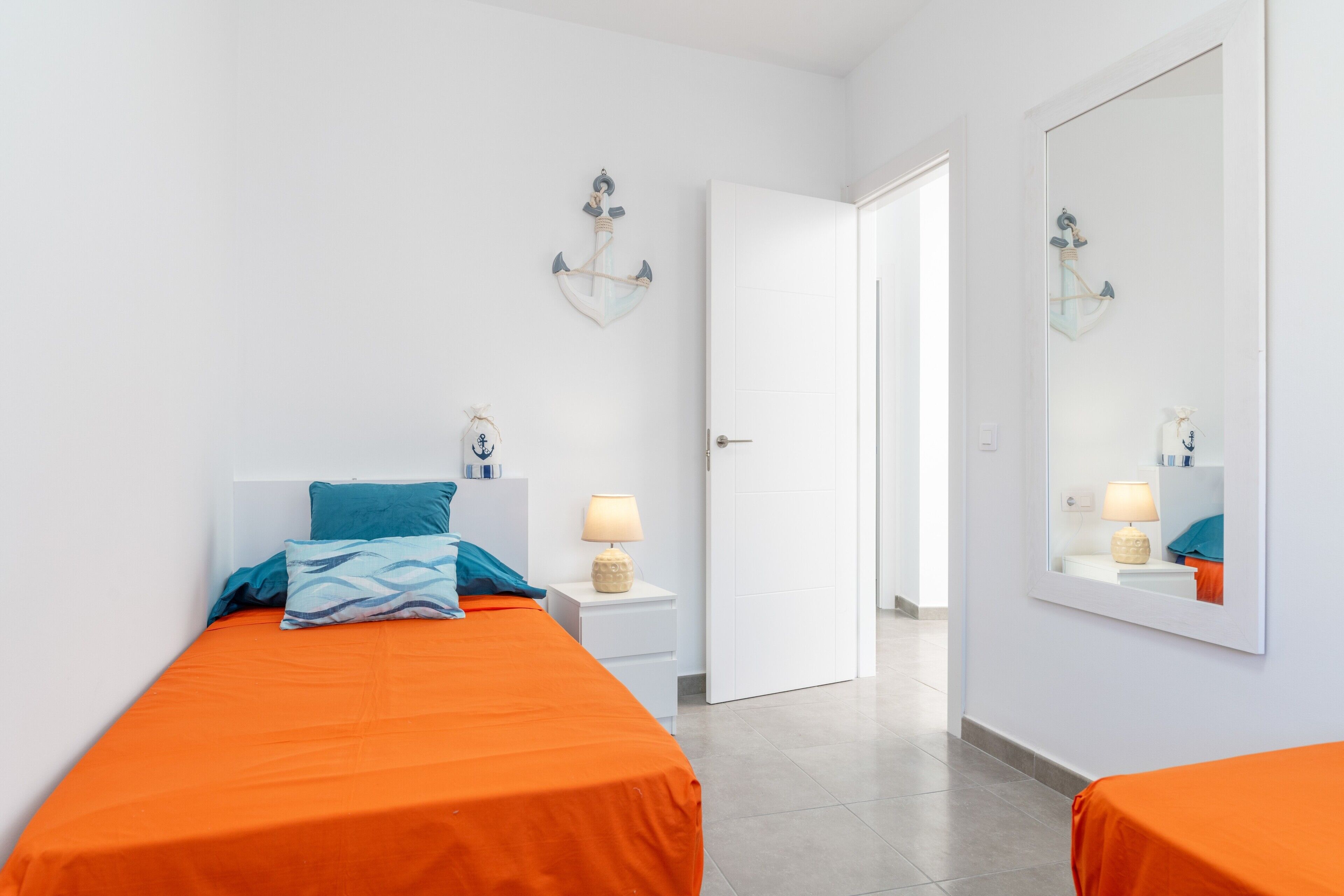 A hotel room with two beds, white walls, a door, a mirror, and a nightstand with a lamp.