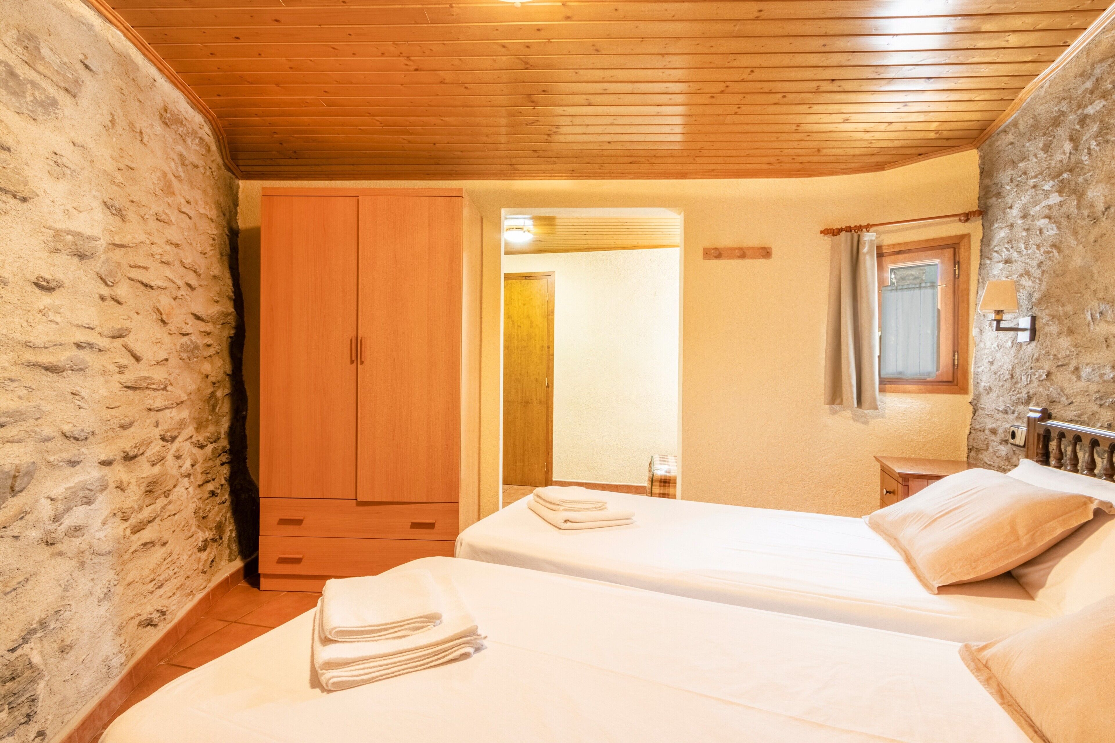 A room with two beds, wooden ceiling, stone walls, a wooden wardrobe, and a small window with a curtain.