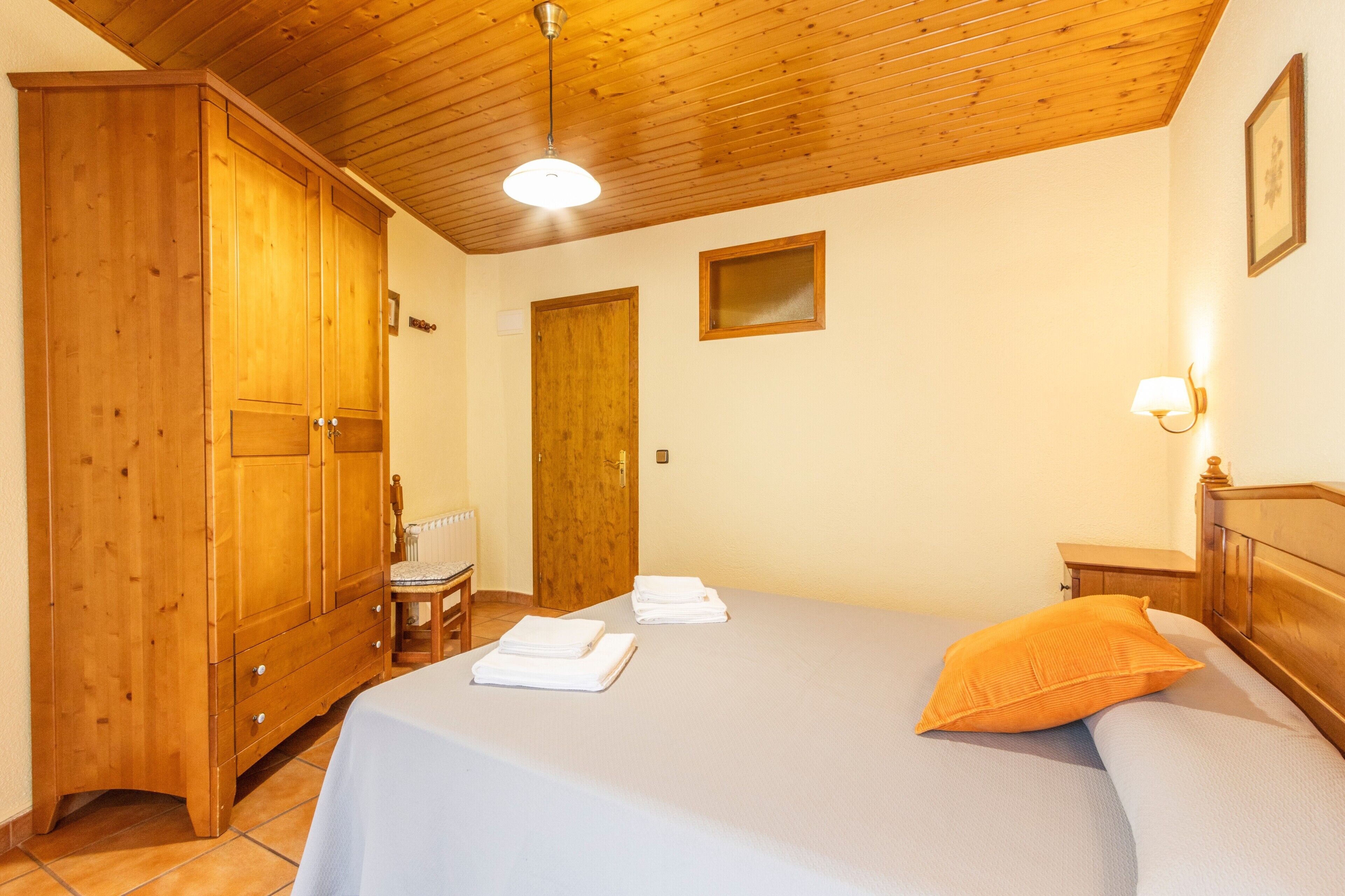 Un dormitorio con un armario de madera, una cama con cubrecama blanco y almohada naranja, una mesita de noche pequeña, y una lámpara de pared.