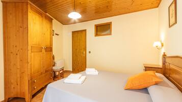 Un dormitorio con un armario de madera, una cama con cubrecama blanco y almohada naranja, una mesita de noche pequeña, y una lámpara de pared.