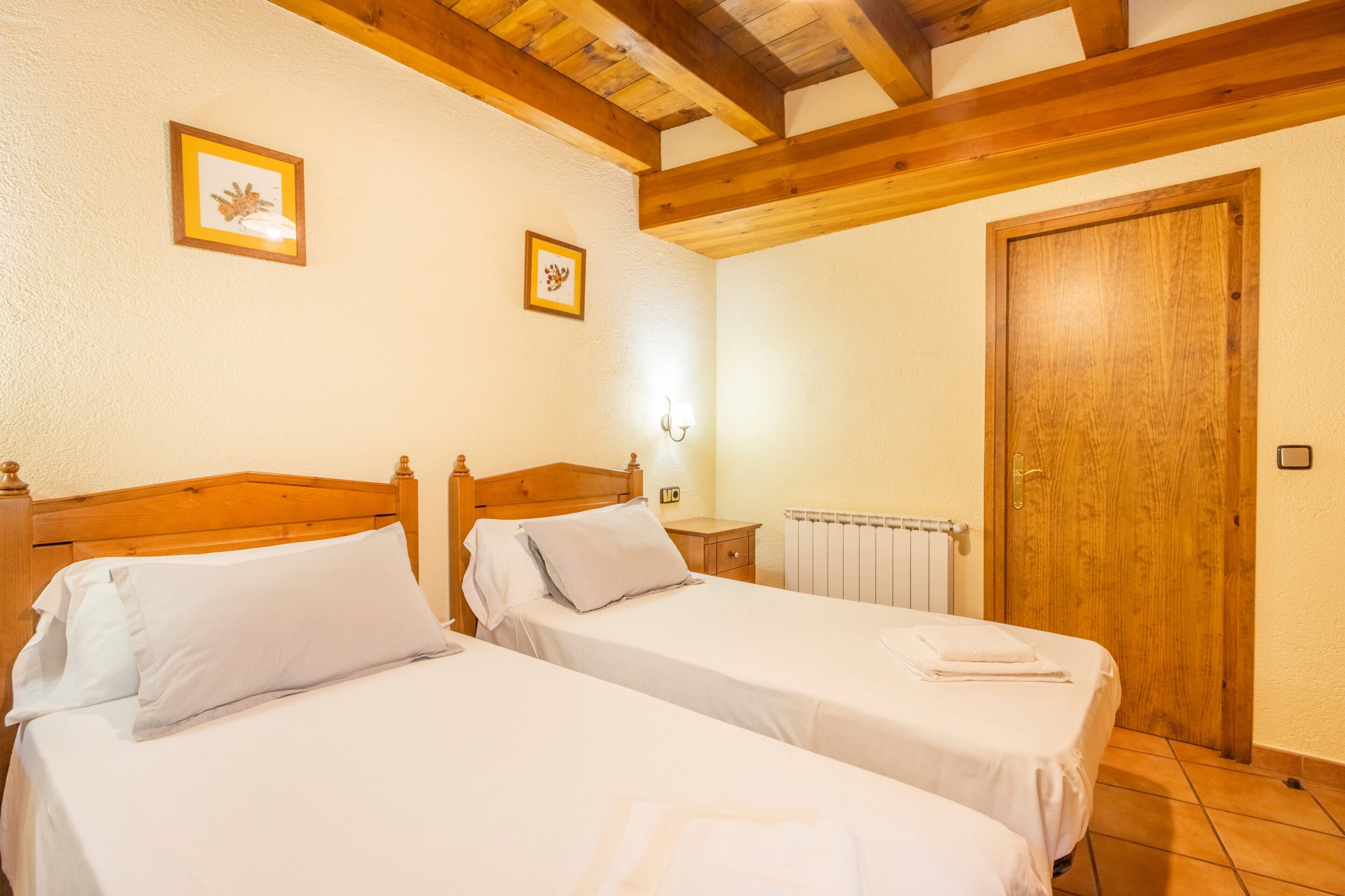 Habitación con dos camas, cabeceros de madera, techo de madera, pared blanca, puerta de madera y una mesita de noche con lámpara.