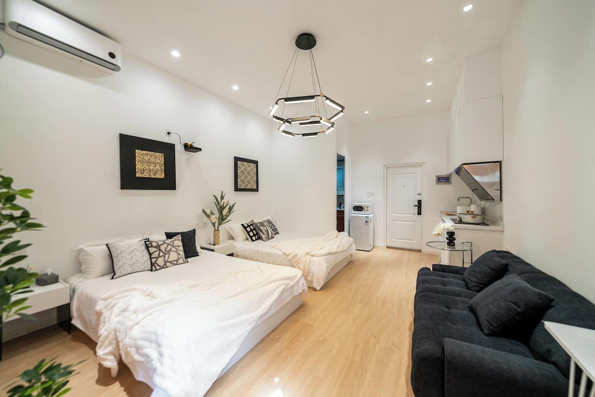 A modern hotel room with two beds, a black sofa, a small kitchen area, and a ceiling-mounted air conditioner.