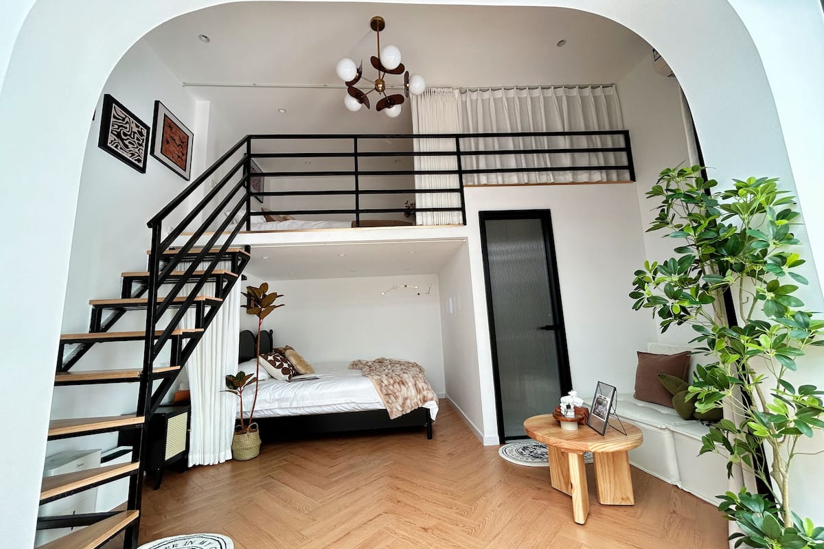 A compact studio apartment with a bunk bed, a staircase with a black railing, a small round wooden table, and a potted plant.