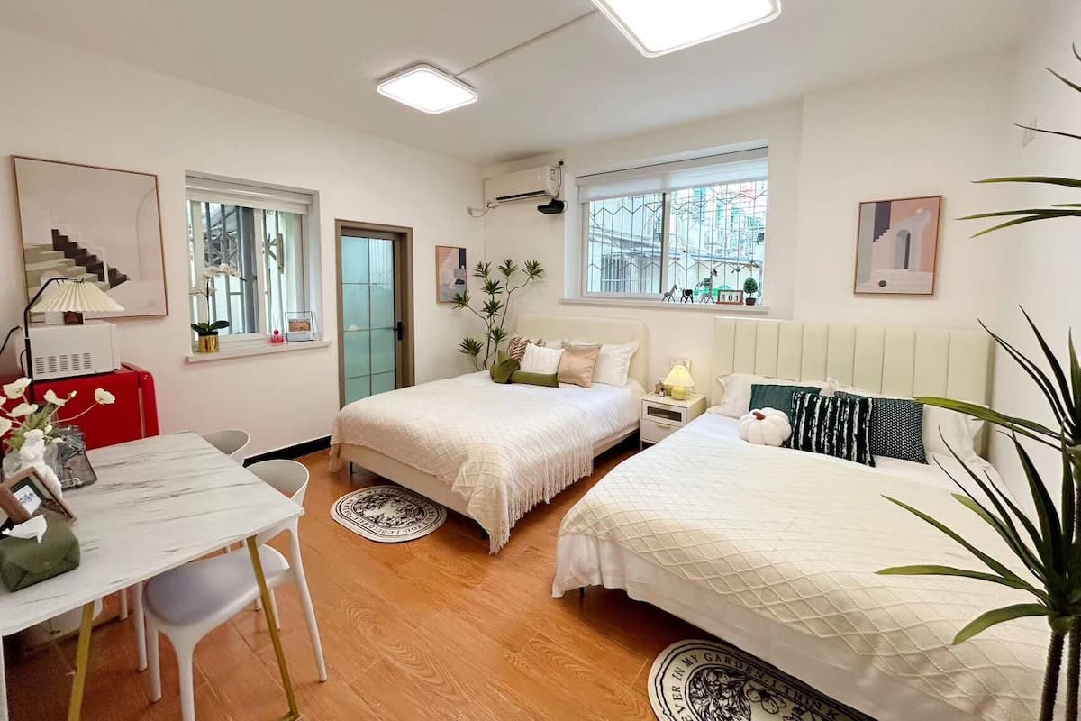 A well-lit, modern bedroom with two beds, a desk, and a chair. There are windows with views of the city, and the room has a clean, minimalist design.