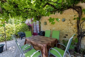 A stone wall with a wooden table and green chairs, surrounded by greenery and a pergola. - Cottage 'Gite Soleil' with Shared Pool, Private Terrace and Wi-Fi (Degagnac)