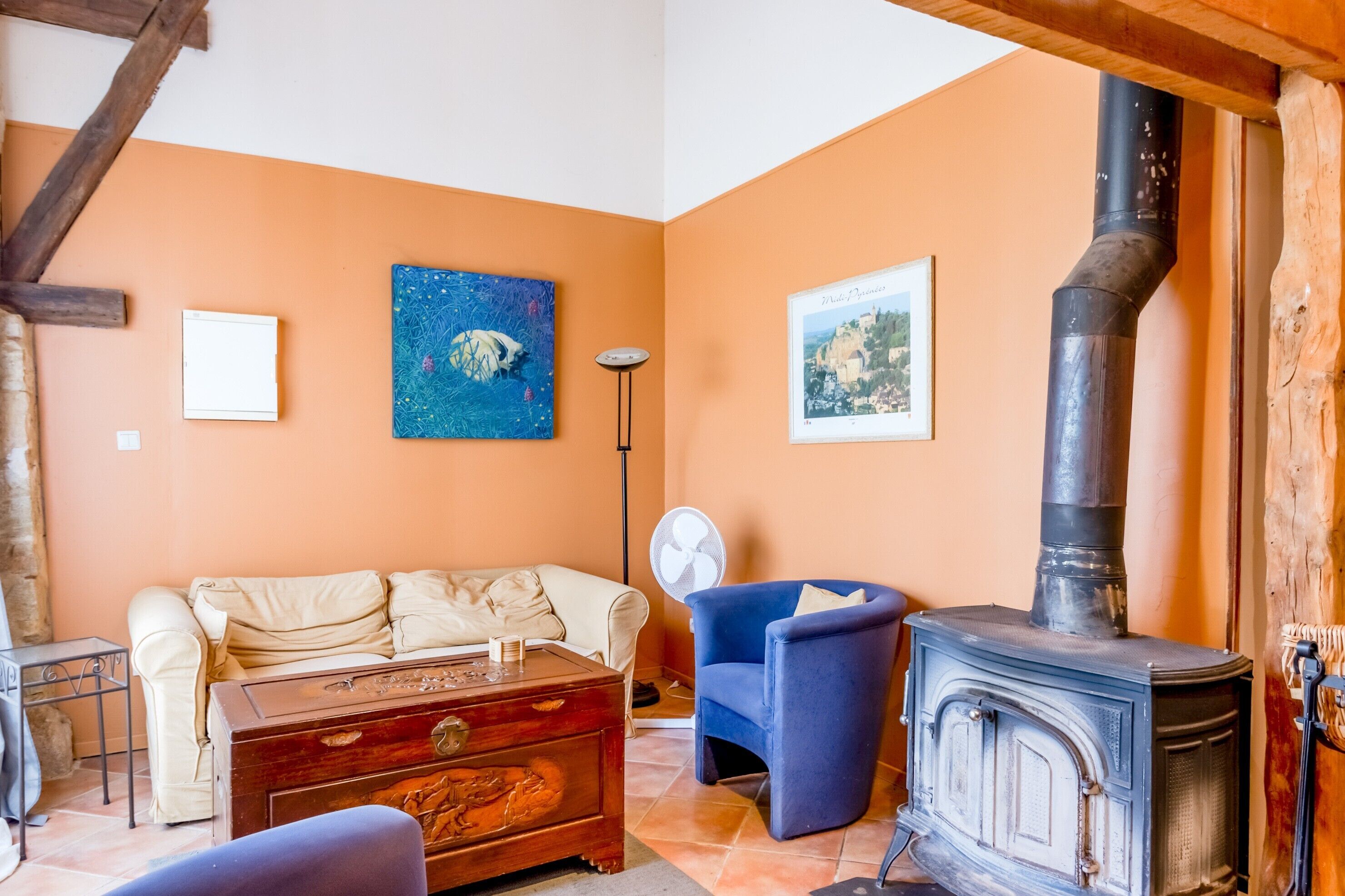 A cozy living room with a sofa, a blue armchair, a wooden chest, a fireplace, and a fan.