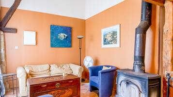 A cozy living room with a sofa, a blue armchair, a wooden chest, a fireplace, and a fan.