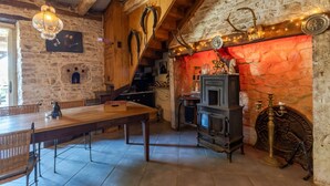 Un intérieur rustique avec une longue table en bois, des murs en pierre, un escalier en bois et un poêle d’époque.