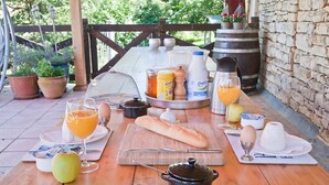 Une table en bois installée à l’extérieur, servant de table pour le petit-déjeuner, avec une miche de pain, des œufs, une casserole et une bouteille de jus.