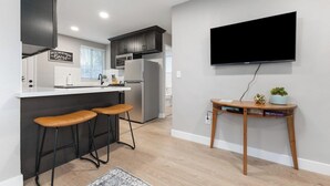 A modern kitchen with dark cabinets, a white countertop, and a built-in oven. There are two bar stools and a small wooden table with a plant and books. A flat-screen TV is mounted on the wall. - The General Sherman Cabin Sequoia Motel (Three Rivers)
