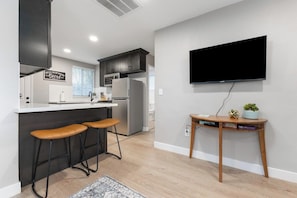 A modern kitchen with dark cabinets, a white countertop, and a built-in oven. There are two bar stools and a small wooden table with a plant and books. A flat-screen TV is mounted on the wall.