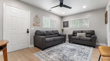 Ein Wohnzimmer mit zwei dunklen Sofas, einem gemusterten Teppich, einem Deckenventilator und einer weißen Tür.