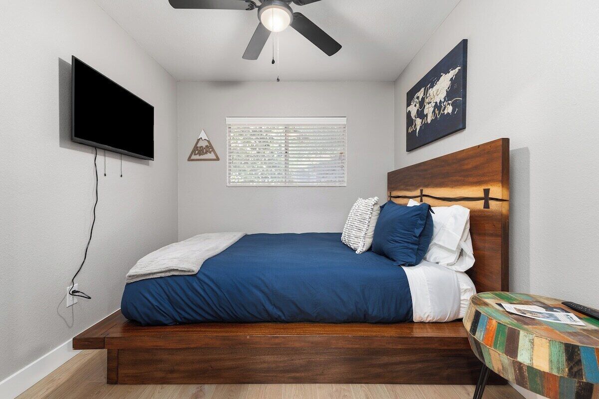 A bedroom with a bed, a ceiling fan, a TV mounted on the wall, a window with blinds, and a small table with a colorful top.