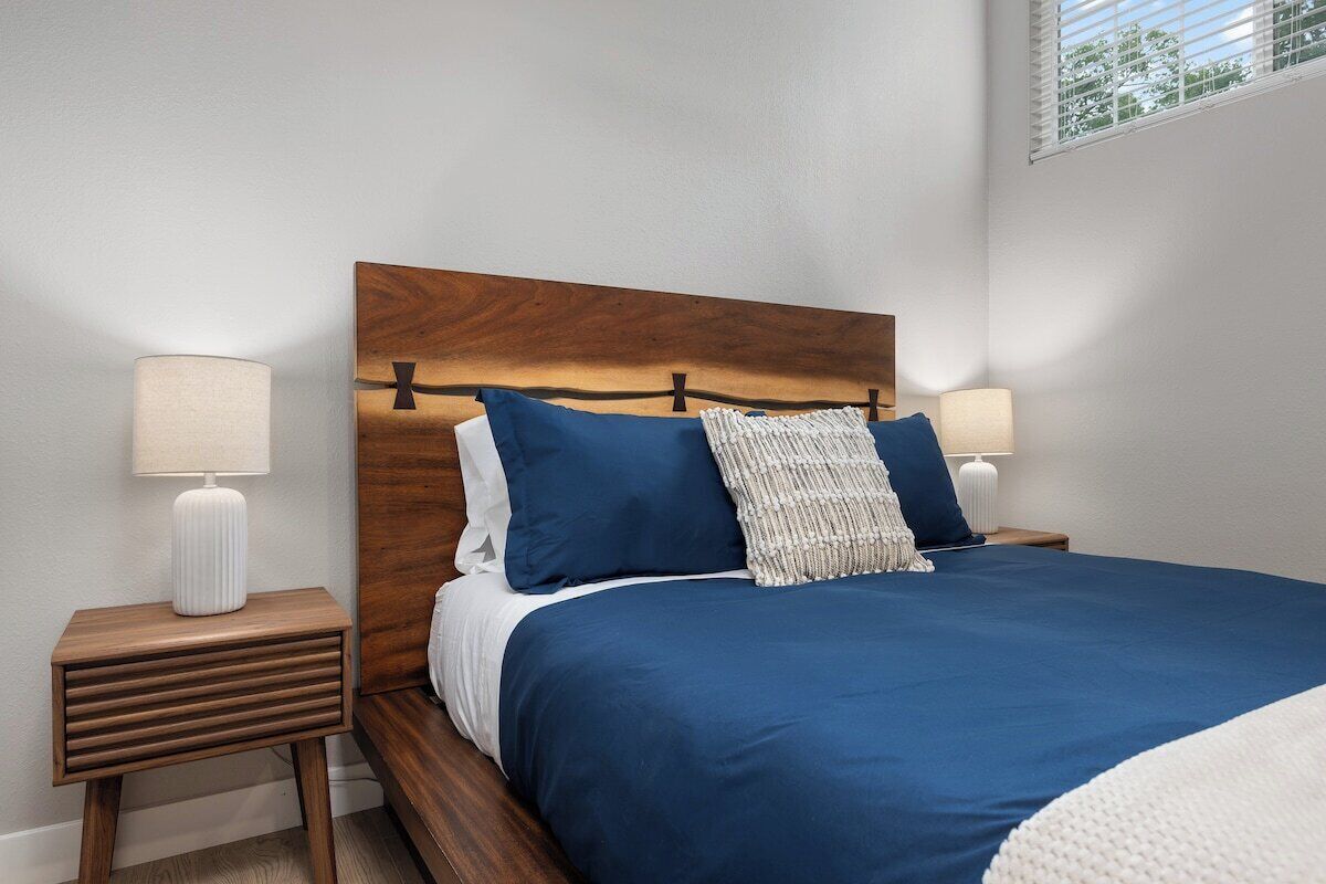 A bedroom with a wooden headboard, two bedside lamps, a blue bedspread, and a white throw blanket.