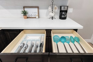 A kitchen counter with a coffee maker, mugs, and a sign that says 'Life Happens. Coffee Gel.' Below, there are two drawers with utensils and kitchen tools.